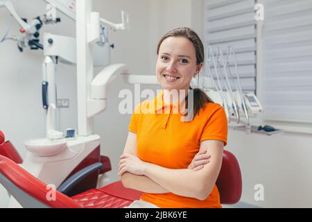 Portrait of a young beautiful female dentist, assistant in orange medical uniform, sitting in the dental office, looking at the camera, arms crossed, Stock Photo