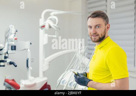 Portrait of a young dentist, assistant in a yellow medical uniform, in a medical black mask and black gloves, with tools in hand, sitting in the denta Stock Photo