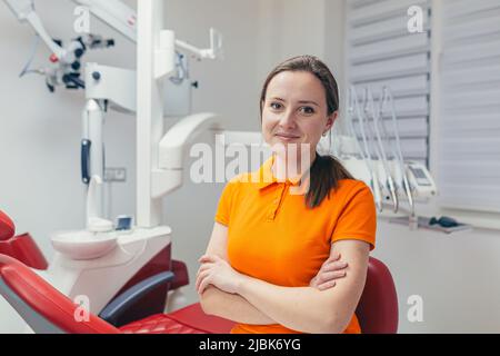 Portrait of a young beautiful female dentist, assistant in orange medical uniform, sitting in the dental office, looking at the camera, arms crossed, Stock Photo