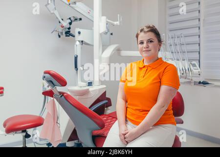 Portrait of a young beautiful female dentist, assistant in orange medical uniform, sitting in the dental office, looking at the camera, arms crossed, Stock Photo