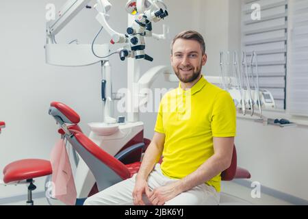 Portrait of a young dentist, assistant in a yellow medical uniform, sitting in a dental office, looking at the camera, arms crossed, smiling Stock Photo