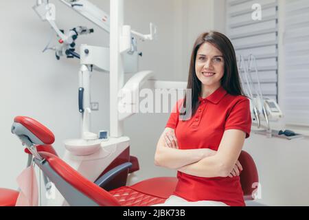 Portrait of a young beautiful female dentist, assistant in a red medical uniform, sitting in the dentist's office, looking at the camera, arms crossed Stock Photo