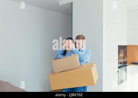 Young couple, man and woman move into a new apartment, house, with boxes and things, surprise each other, close their eyes, smile Stock Photo