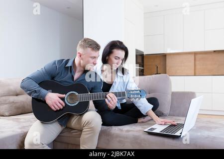 Young couple, man and woman at home on the couch playing guitar online from laptop, studying, giving lessons, kissing, smiling Stock Photo