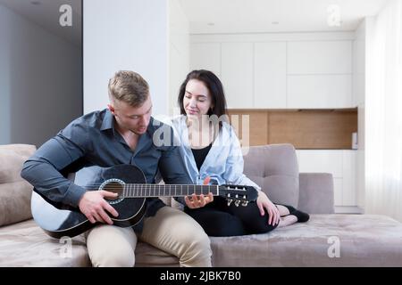 Young couple, man and woman at home on the couch playing guitar online from laptop, studying, giving lessons, kissing, smiling Stock Photo