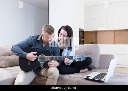 Young couple, man and woman at home on the couch playing guitar online from laptop, studying, giving lessons, kissing, smiling Stock Photo