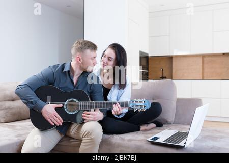 Young couple, man and woman at home on the couch playing guitar online from laptop, studying, giving lessons, kissing, smiling Stock Photo