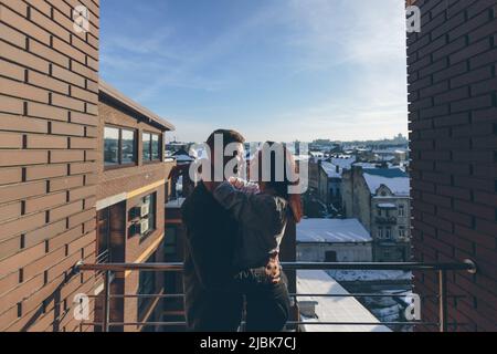 Loving beautiful young couple, man and woman standing on the balcony on the terrace of the house, sunlight, hugging kissing Stock Photo
