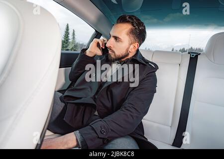 Caucasian young successful man seriously talking on the smartphone while sitting in the back seat of a driving car Stock Photo