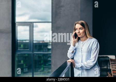 Beautiful blonde woman uses a mobile phone while standing on the balcony of an apartment building Stock Photo