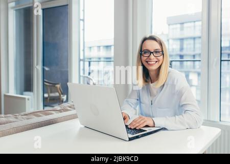 Beautiful young woman with glasses working at home on laptop, online, studying, teaching, making a video call, student, looking at camera, smiling Stock Photo