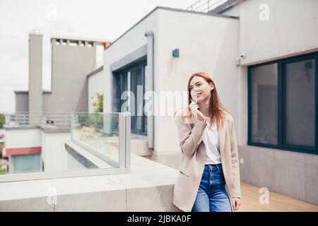 young caucasian woman standing on the terrace of a modern office or balcony at home and smokes marijuana. Business lady smoking cannabis. female relie Stock Photo