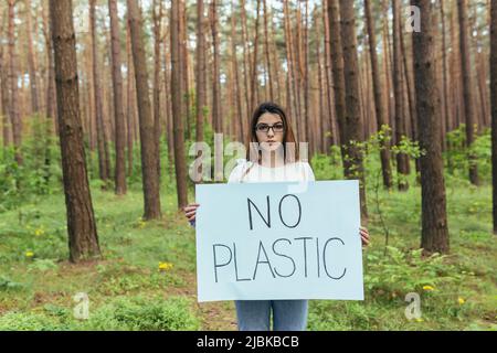 Young female activist standing in the woods with no plastic poster, volunteer struggling with forest pollution Stock Photo