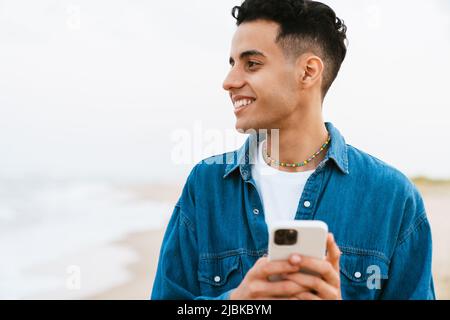Young middle eastern man smiling while using cellphone at summer beach Stock Photo