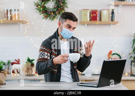 A sick man with a cold in a protective mask works remotely from home using a laptop during the New Year and Christmas holidays communicates with colle Stock Photo