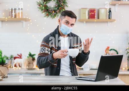 A sick man with a cold in a protective mask works remotely from home using a laptop during the New Year and Christmas holidays communicates with colle Stock Photo