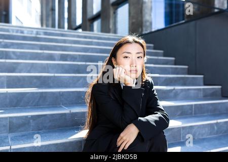 Young beautiful asian woman upset and sad sitting on the stairs of the office center, depressed business woman fired Stock Photo