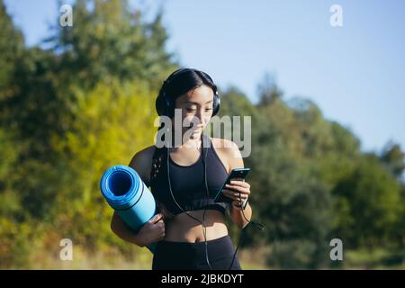 Beautiful asian woman athlete goes to fitness class, listens to music on headphones and uses the phone Stock Photo