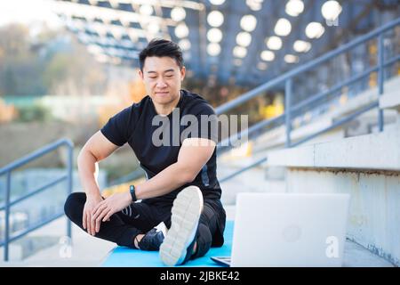 handsome asian fitness coach holds workout classes online using video call webcam and laptop. sitting outside in a modern stadium. Pilates training or Stock Photo
