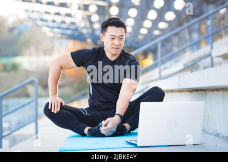 handsome asian fitness coach holds workout classes online using video call webcam and laptop. sitting outside in a modern stadium. Pilates training or Stock Photo