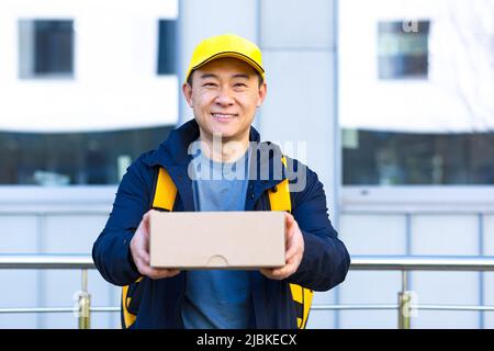 portrait asian courier. happy male delivery service worker in cap and with backpack food bag. man Employee with a box or parcel in hand on a street lo Stock Photo