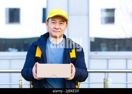 Walking courier in protective medical mask on face delivery man asian smiling looking for delivery address on phone, has big yellow backpack for food Stock Photo