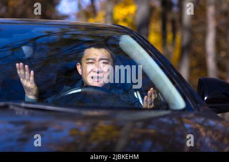 Furious angry asian man standing in a traffic jam beeps and hurries. Sitting in the car. Irritated young business male tired driver in automobile. Str Stock Photo
