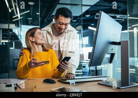 Creative team of Asian man and woman shopping online, sitting at a table, choosing a gift together, rejoicing and smiling, using a smartphone and cred Stock Photo