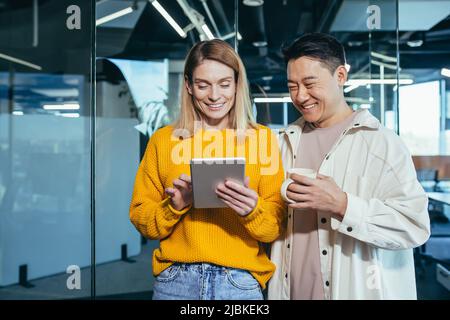Two fellow programmers, a man and a woman, happy and smiling looking at the tablet screen, working in a modern office, lunch break Stock Photo