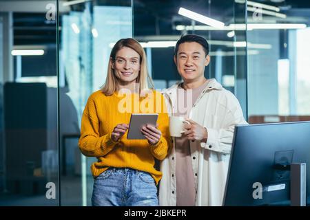 a team of two Asian male and female workers take a break, look at the camera and smile, use a tablet to discuss a joint business project Stock Photo