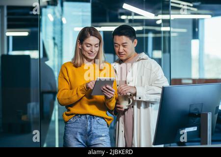 Two fellow programmers, a man and a woman, happy and smiling looking at the tablet screen, working in a modern office, lunch break Stock Photo
