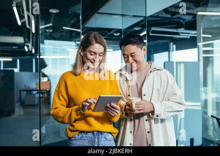 Two fellow programmers, a man and a woman, happy and smiling looking at the tablet screen, working in a modern office, lunch break Stock Photo