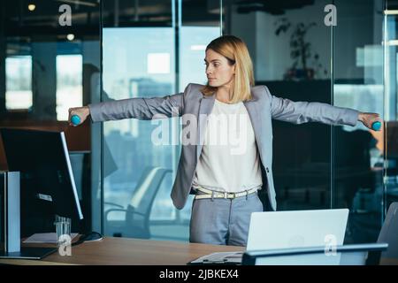 Beautiful and happy woman doing sports exercises in the office at work, during a break, business woman monitors health engaged in fitness Stock Photo