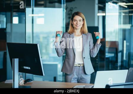 Beautiful and happy woman doing sports exercises in the office at work, during a break, business woman monitors health engaged in fitness Stock Photo