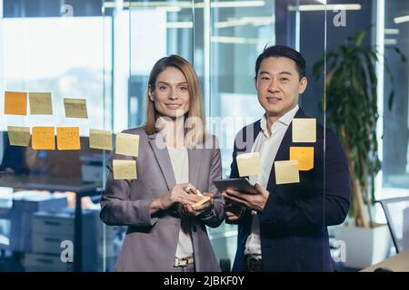Startup and implementation of a small business project. Asian Businessman discussing with businesswoman sticking adhesive notes on glass wall in offic Stock Photo