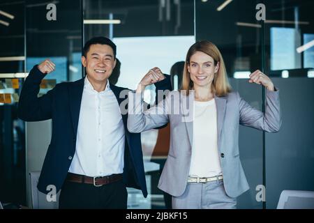 A team of successful managers, an Asian man and a woman, in business attire look at the camera and smile hold hands up, gesture of strength, and can p Stock Photo