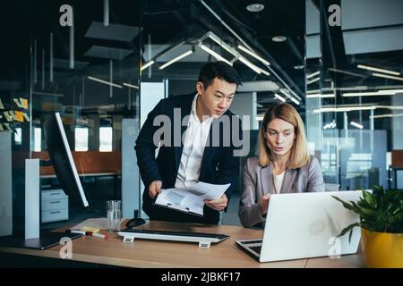Two employees in a modern office, an Asian man and a woman working at a table, colleagues discussing and consulting, thinking about a joint project Stock Photo
