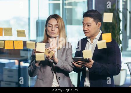 Startup and implementation of a small business project. Asian Businessman discussing with businesswoman sticking adhesive notes on glass wall in offic Stock Photo