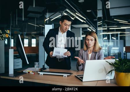 Two employees in a modern office, an Asian man and a woman working at a table, colleagues discussing and consulting, thinking about a joint project Stock Photo