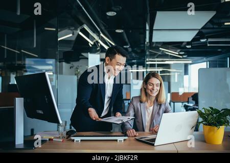 Two employees in a modern office, an Asian man and a woman working at a table, colleagues discussing and consulting, thinking about a joint project Stock Photo