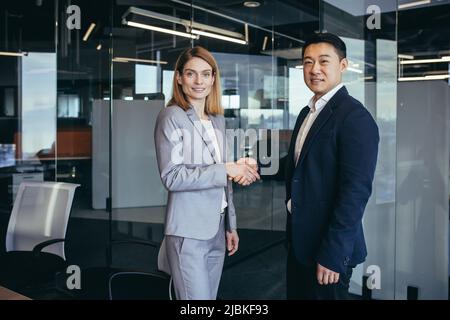 Asian businessman shakes hands with business woman employee, happy team of workers looking at camera and smiling, successful conclusion of negotiation Stock Photo