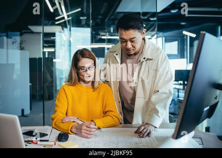Two employees of designers and architects, thinking about the project, an Asian man and a blonde woman, in casual clothes working together at the tabl Stock Photo