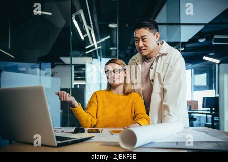 Two employees of designers and architects, thinking about the project, an Asian man and a blonde woman, in casual clothes working together at the tabl Stock Photo