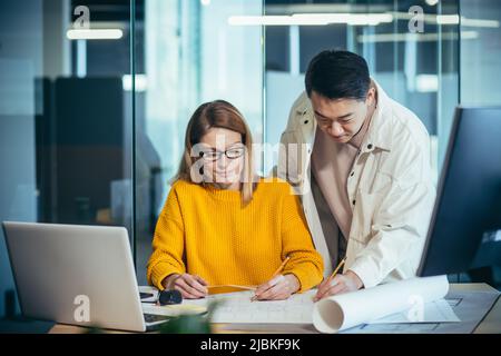 Two employees of designers and architects, thinking about the project, an Asian man and a blonde woman, in casual clothes working together at the tabl Stock Photo
