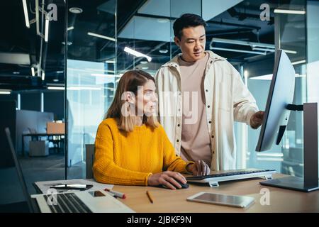 Two freelancers, an Asian man and a woman, work in a modern office, discussing and discussing a joint project, looking at a computer monitor Stock Photo