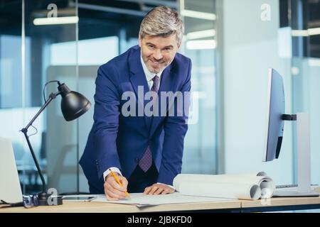 Experienced gray-haired designer architect, smiling and looking at camera, man working on architectural projector of new office center Stock Photo