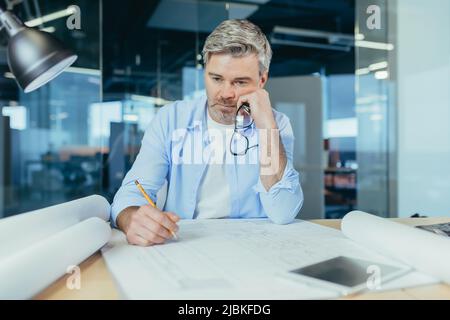 Thinking senior male architect working on an important project. at the table with a computer and a drawing Stock Photo