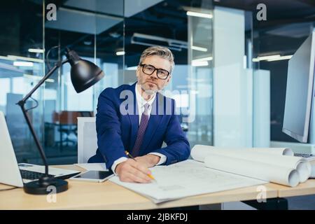 Experienced gray-haired designer architect, smiling and looking at camera, man working on architectural projector of new office center Stock Photo