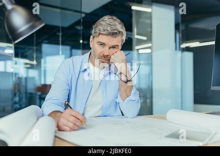 Thinking senior male architect working on an important project. at the table with a computer and a drawing Stock Photo