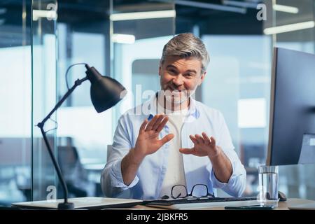Successful businessman works at the computer, sits at the table and looks at the camera, communicates, explains and gestures Stock Photo